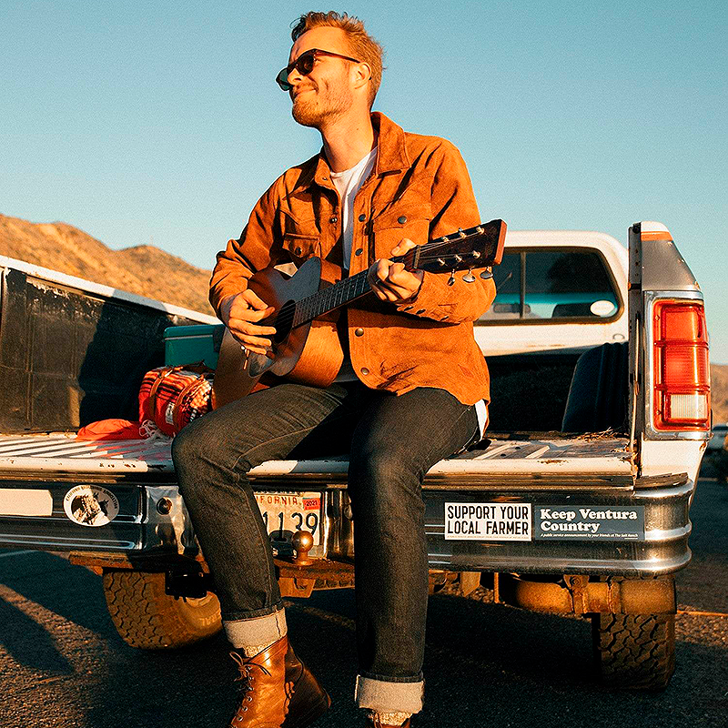 Man playing guitar in the bed of a pickup truck with mountains in the background