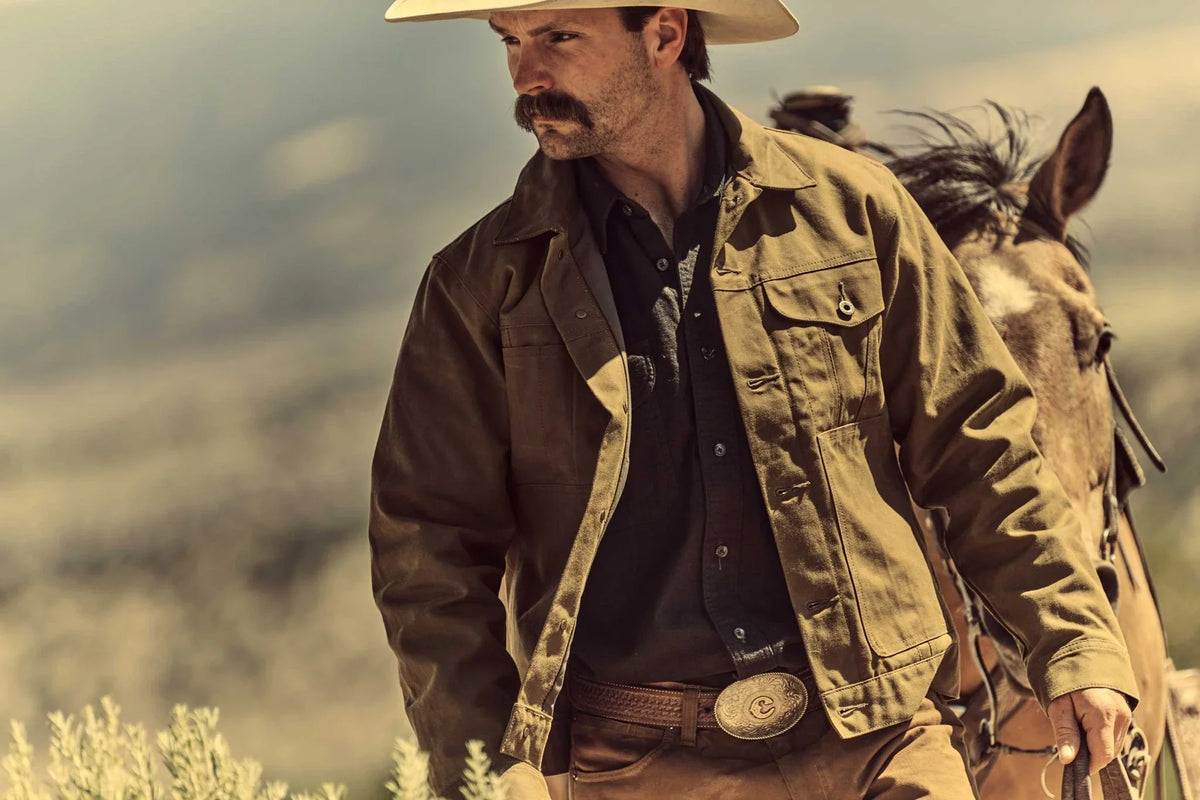 A man in a cowboy hat and brown jacket walking beside a horse in an outdoor, blurred landscape.