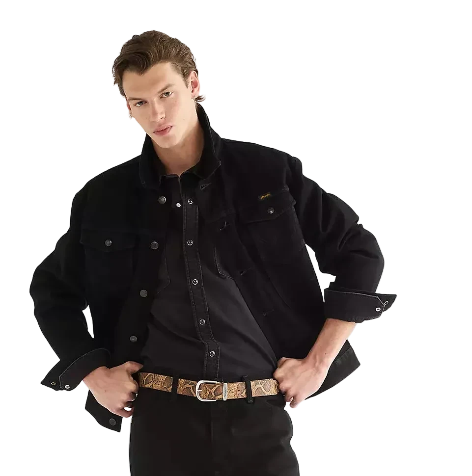 A male model wearing a black Wrangler Cowboy Cut denim jacket over a black button-down shirt, black jeans, a snake patterned belt, and brown suede boots.