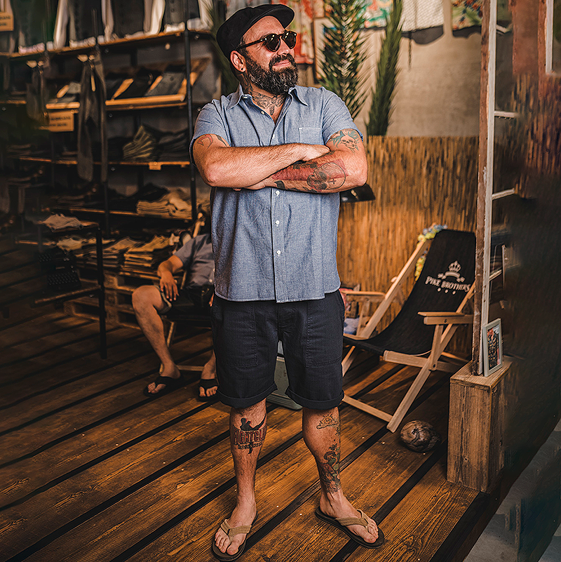 Man with tattoos standing in a rustic indoor setting with wooden floors and furniture.
