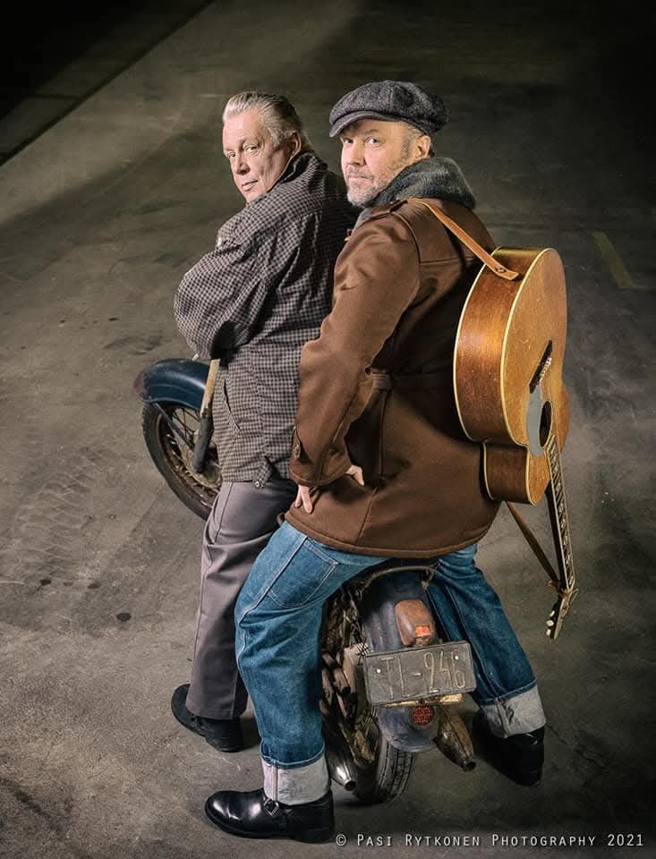 Two men with a guitar and motorcycle in an urban setting
