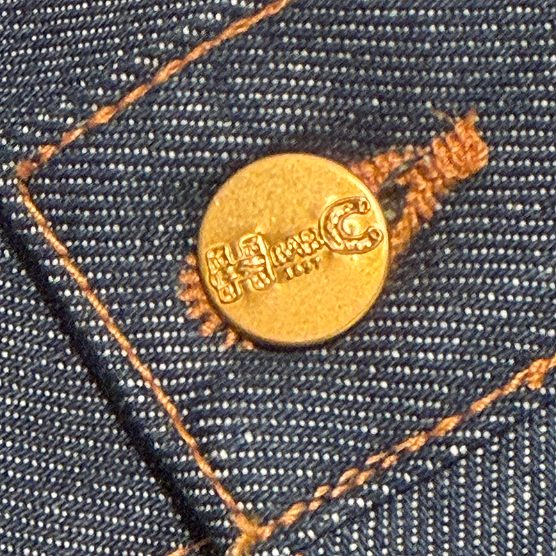 Close-up of a brass button with a H Bar C logo on a denim fabric background