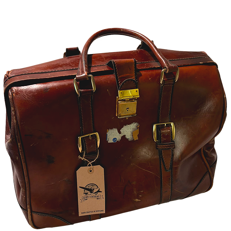 A vintage brown leather duffel bag with gold-tone hardware, featuring a top handle, buckle straps, a hanging tag, and a small peeling sticker on the front.