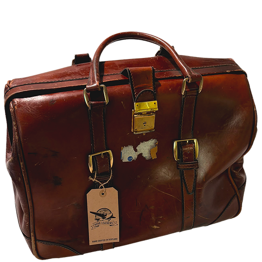 A vintage brown leather duffel bag with gold-tone hardware, featuring a top handle, buckle straps, a hanging tag, and a small peeling sticker on the front.