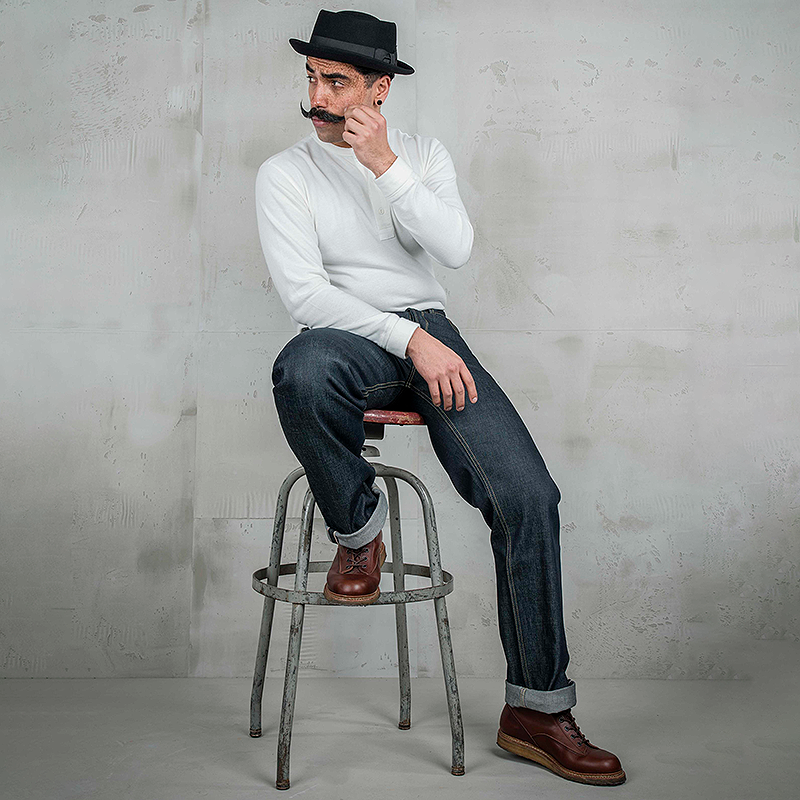 A man wearing a white long-sleeve shirt, dark blue jeans, brown leather shoes, and a black hat sits on a metal stool against a textured gray wall.