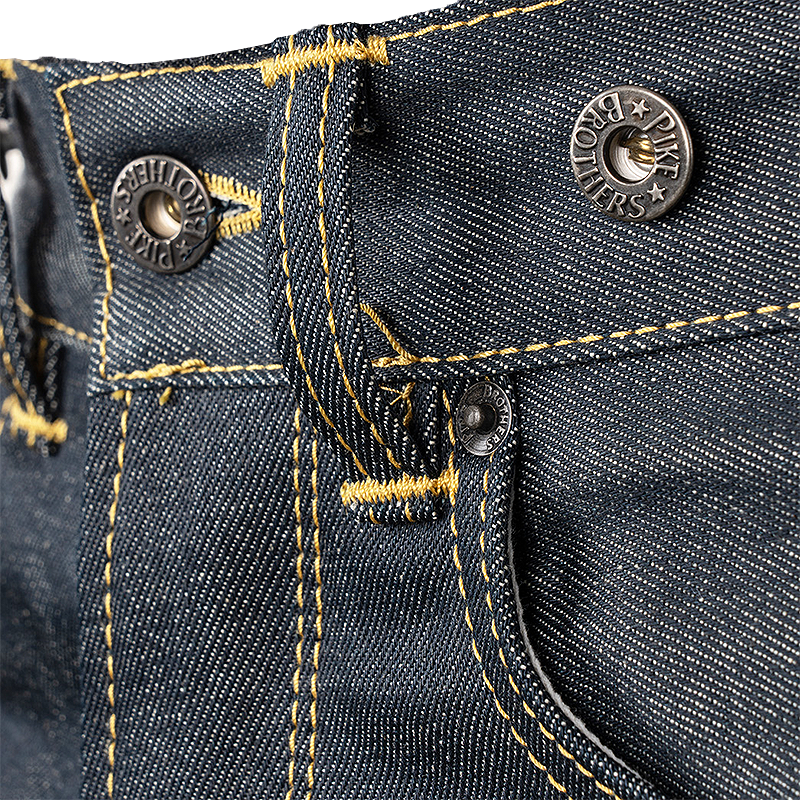 Close-up of dark indigo denim jeans showing the waistband, yellow contrast stitching, metal rivets, and a buttons with Pike Brothers branding.