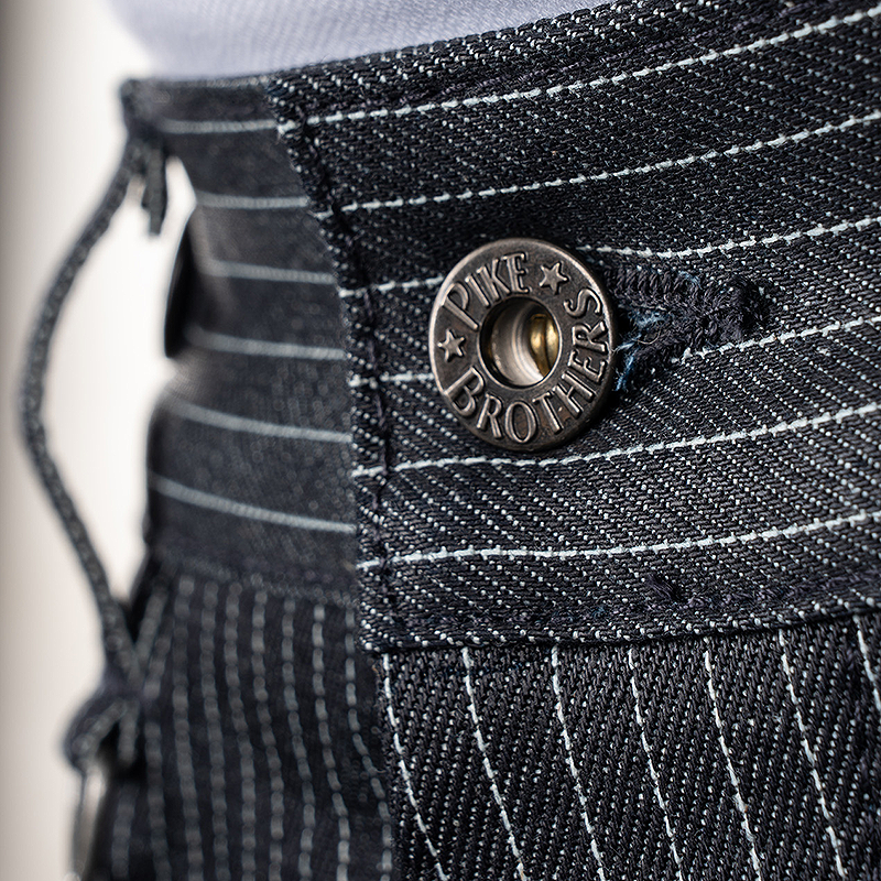 Close-up of an indigo blue pinstriped denim garment, showing a round metal button engraved with 'PIKE BROTHERS'.