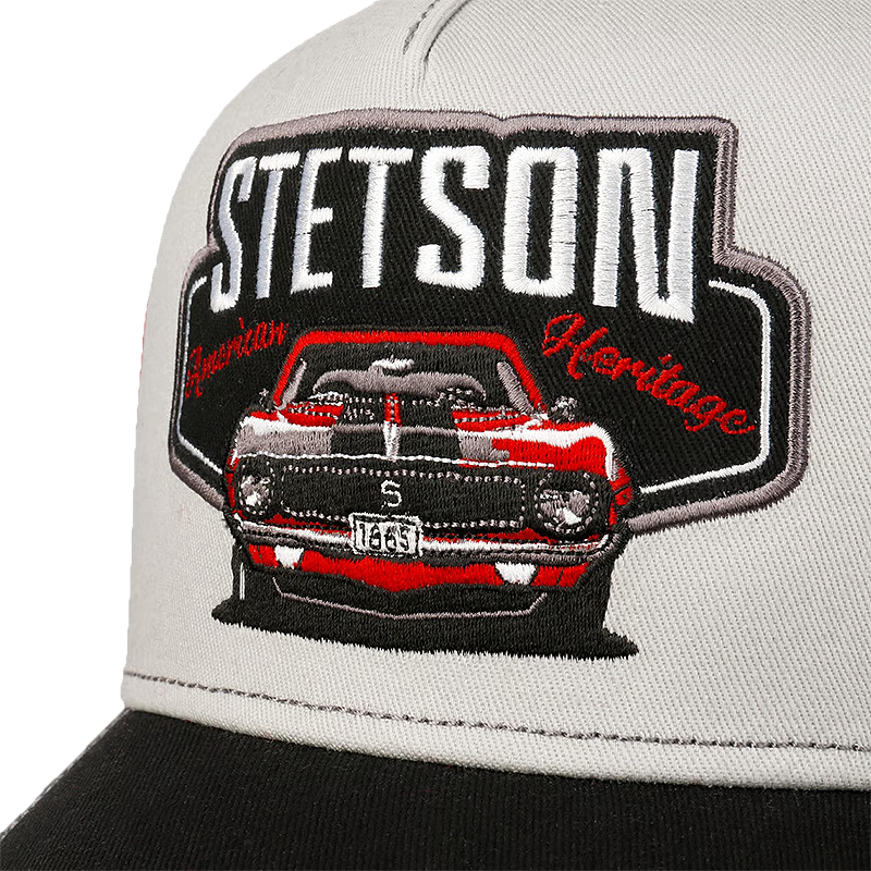Close-up of Stetson Garage American Heritage Trucker Cap with a black brim, featuring a large embroidered patch on the front. The patch displays a red and gray muscle car with the text 'STETSON American Heritage' in white and red.