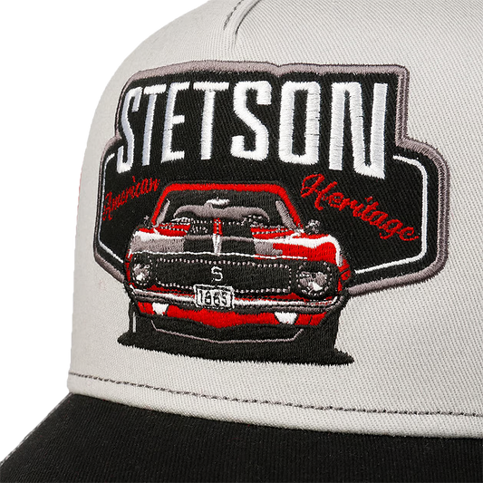 Close-up of Stetson Garage American Heritage Trucker Cap with a black brim, featuring a large embroidered patch on the front. The patch displays a red and gray muscle car with the text 'STETSON American Heritage' in white and red.