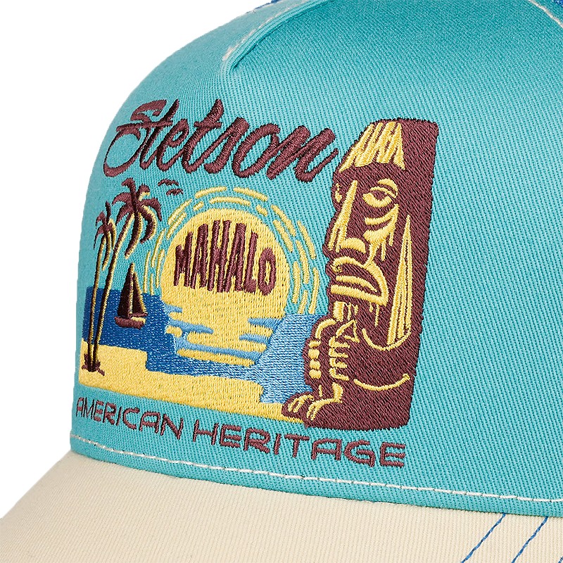 Close-up of a turquoise Stetson Mahalo Trucker Cap with a detailed embroidered graphic on the front panel. The embroidery features a tiki figure, palm trees, a sailboat, a yellow sun, the words 'Stetson' and 'MAHALO', and 'AMERICAN HERITAGE' in maroon.