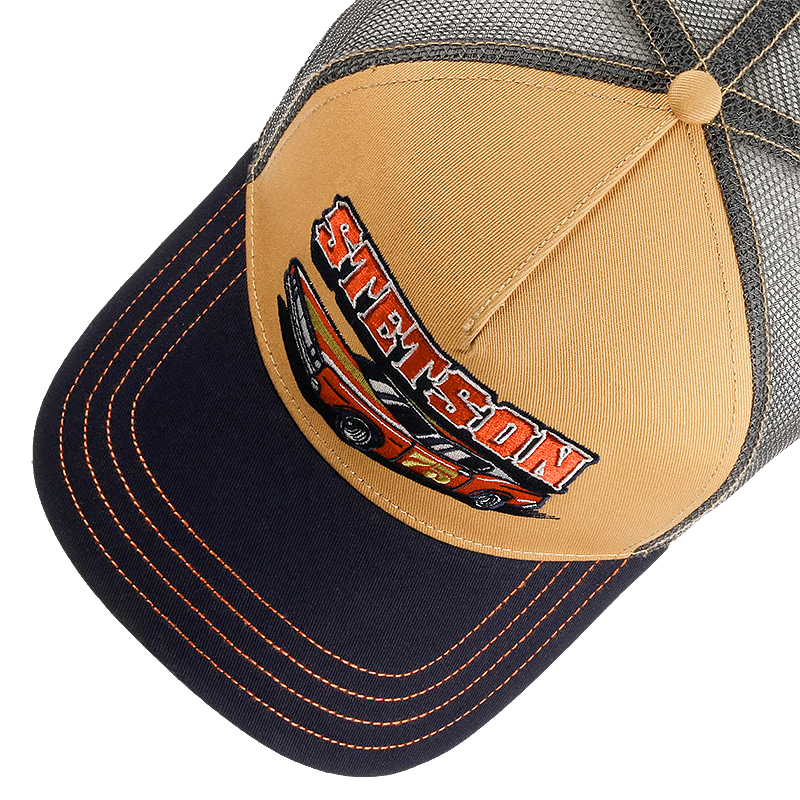 A close-up of a trucker-style baseball cap with a tan front panel, dark navy brim, and gray mesh back. The front panel features a large embroidered patch with the name 'STETSON' in red and orange outlined letters above a red, orange, and black race car.