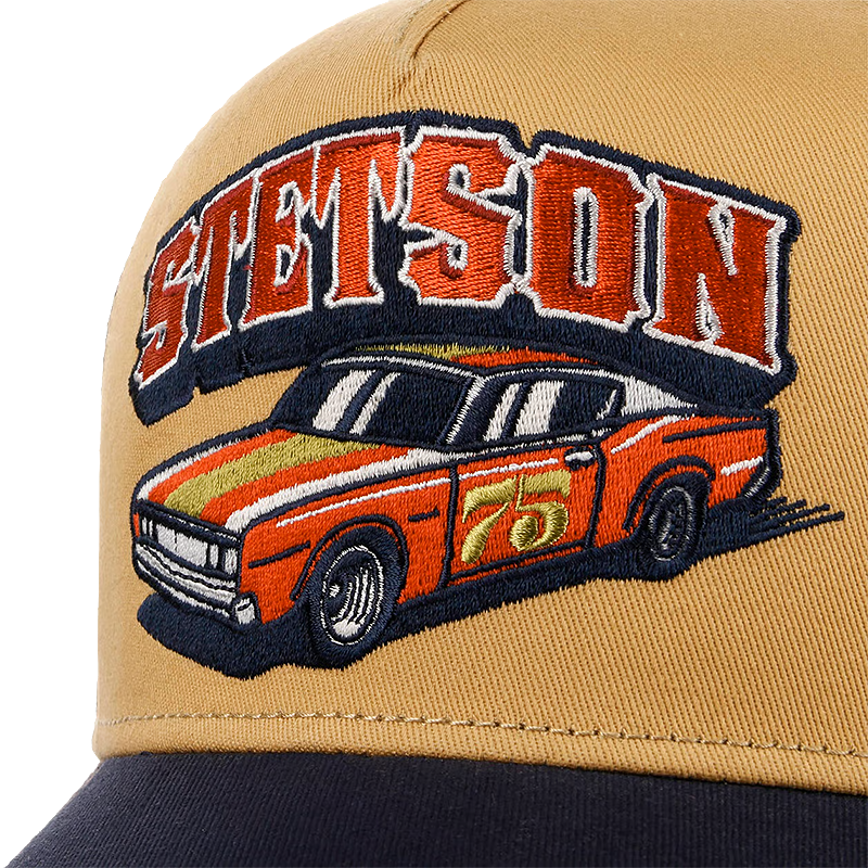 Close-up of a tan Stetson Indy Trucker Cap with a large embroidered patch featuring the word 'STETSON' in red and white outlined letters above an orange race car with a gold stripe and the number 75. The brim of the cap is dark navy.