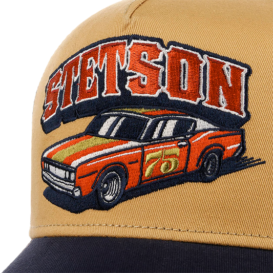 Close-up of a tan Stetson Indy Trucker Cap with a large embroidered patch featuring the word 'STETSON' in red and white outlined letters above an orange race car with a gold stripe and the number 75. The brim of the cap is dark navy.