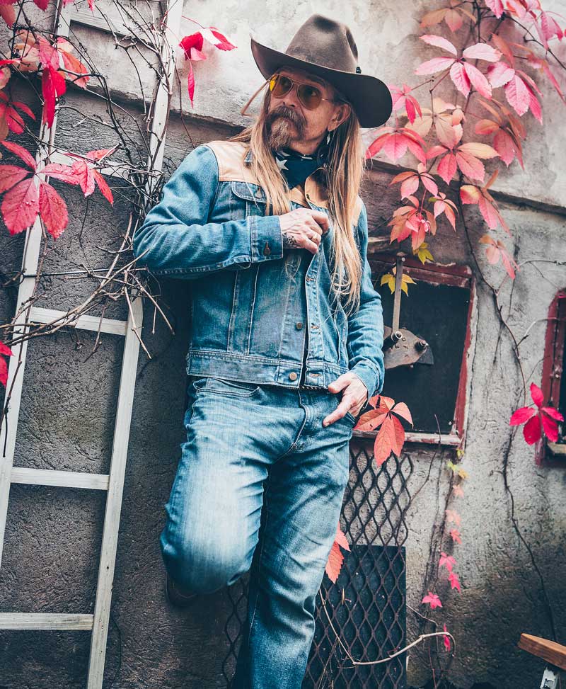 Man standing by a wall wearing denim jacket and jeans.