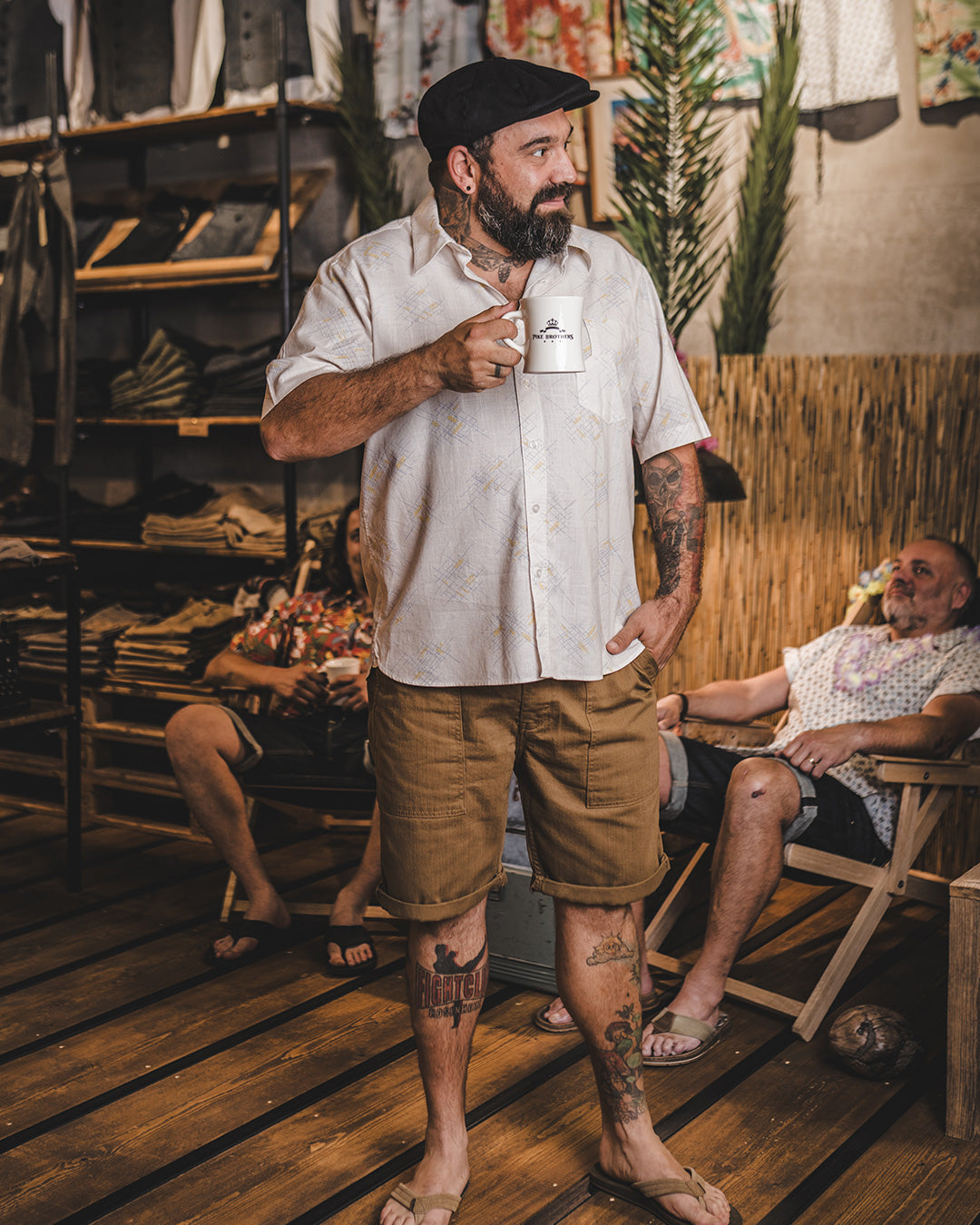 Man holding a coffee cup in a casual indoor setting with wooden floors and people in the background.